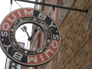 A donut shaped sign with the copy "Quimby's" wrapped twice around hangs down from a brick buildings