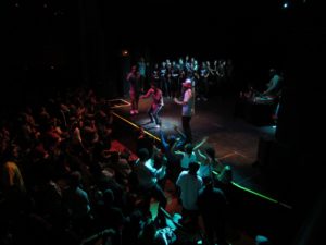 A crowd of people surround a stage with performers in a dark room. 
