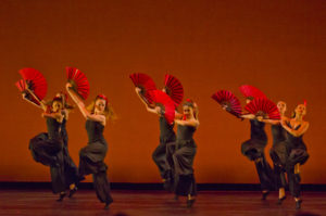 Six ballet dancers dressed in black dressed hold bright red fans in the middle of a dance number.